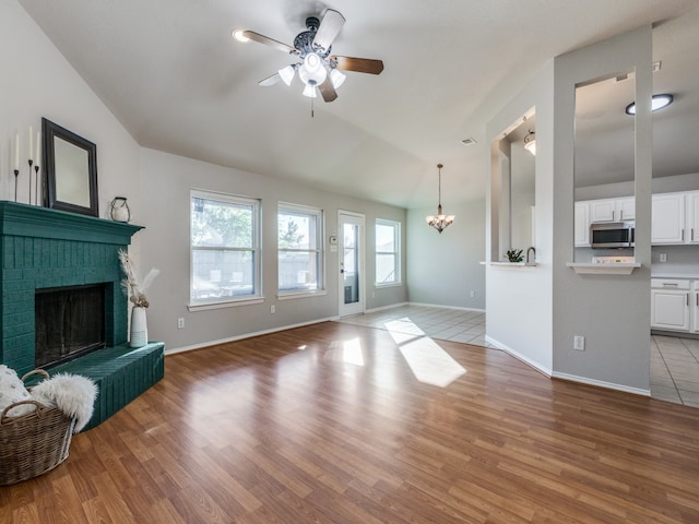 living room with a brick fireplace, ceiling fan with notable chandelier, vaulted ceiling, and light wood-type flooring