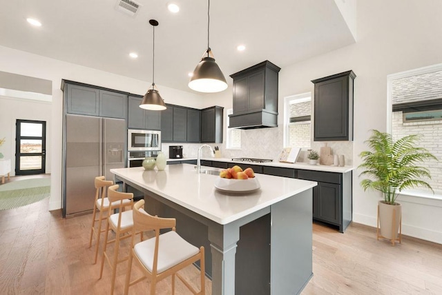 kitchen with pendant lighting, custom exhaust hood, a center island with sink, light hardwood / wood-style flooring, and built in appliances