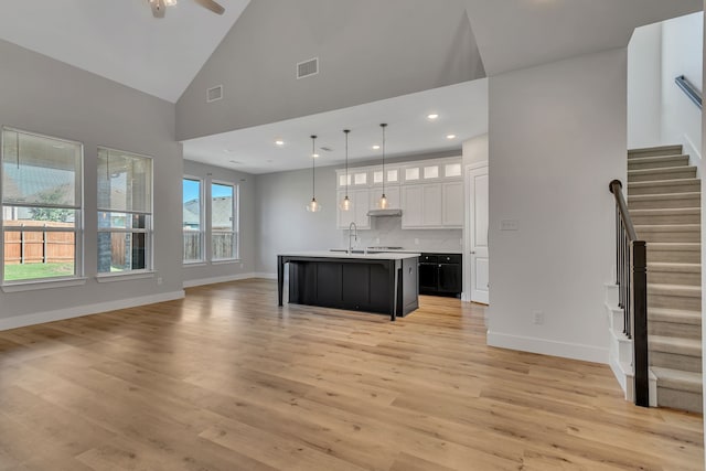 kitchen featuring decorative light fixtures, high vaulted ceiling, a center island with sink, light hardwood / wood-style floors, and white cabinets