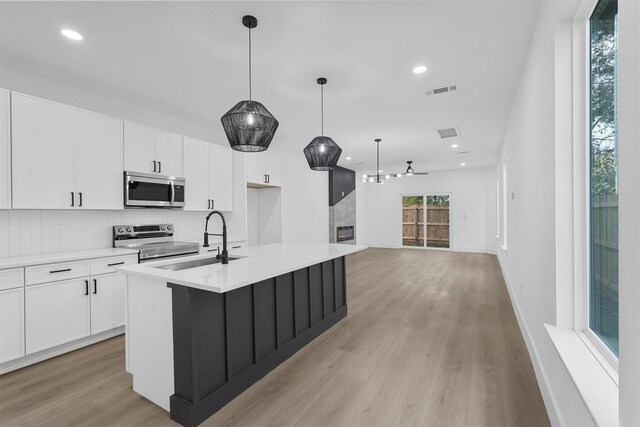 kitchen with stainless steel appliances, a center island with sink, white cabinetry, light wood-type flooring, and decorative light fixtures