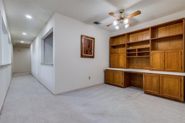 unfurnished office featuring a ceiling fan, visible vents, built in desk, a textured ceiling, and light carpet