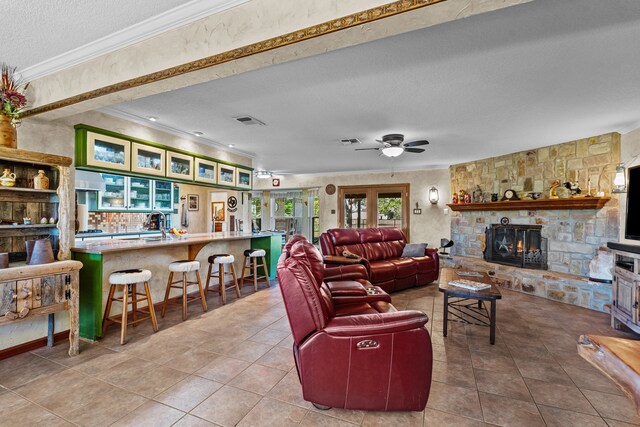 living room with light tile patterned flooring, ceiling fan, a textured ceiling, a stone fireplace, and crown molding