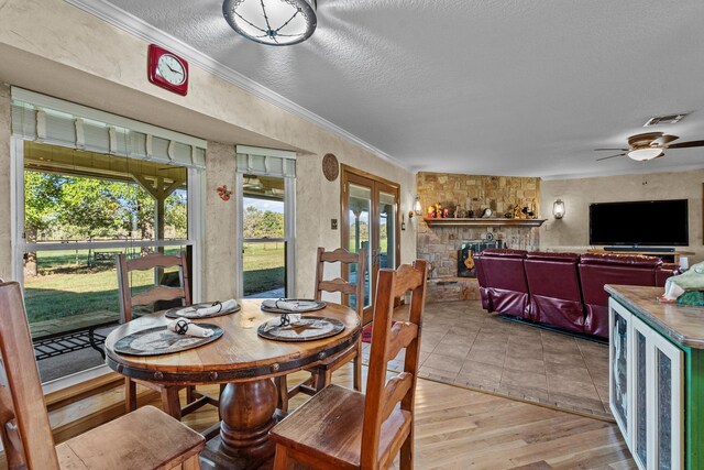 dining area featuring crown molding, a stone fireplace, a textured ceiling, light hardwood / wood-style floors, and ceiling fan