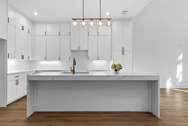 kitchen with light stone counters, sink, white cabinetry, hanging light fixtures, and an island with sink