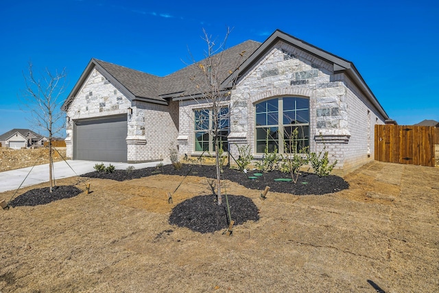 french country style house with an attached garage, brick siding, fence, driveway, and roof with shingles