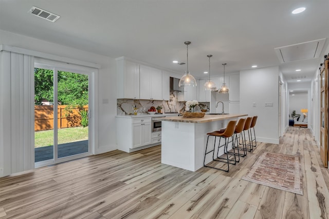 kitchen with light wood-type flooring, wall chimney range hood, decorative light fixtures, white cabinets, and a kitchen island with sink