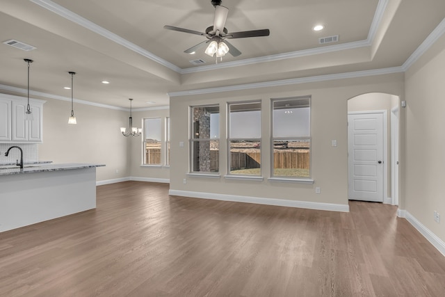 unfurnished living room with ceiling fan with notable chandelier, light wood-type flooring, a raised ceiling, and crown molding