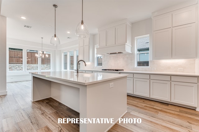 kitchen with light wood-type flooring, hanging light fixtures, an island with sink, and white cabinets