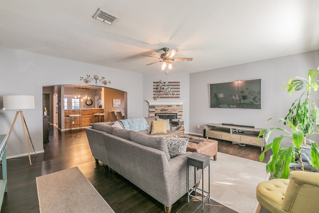 living room with ceiling fan with notable chandelier, a stone fireplace, and dark wood-type flooring