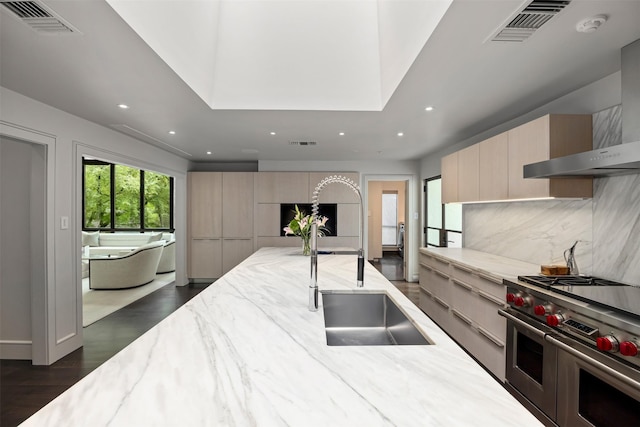 kitchen with light stone countertops, light brown cabinets, wall chimney range hood, backsplash, and range with two ovens