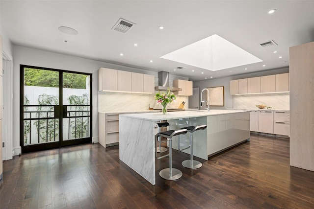 kitchen featuring dark hardwood / wood-style flooring, backsplash, a skylight, a kitchen island with sink, and wall chimney range hood