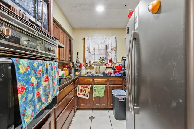 kitchen featuring black oven, light tile patterned floors, sink, and stainless steel refrigerator