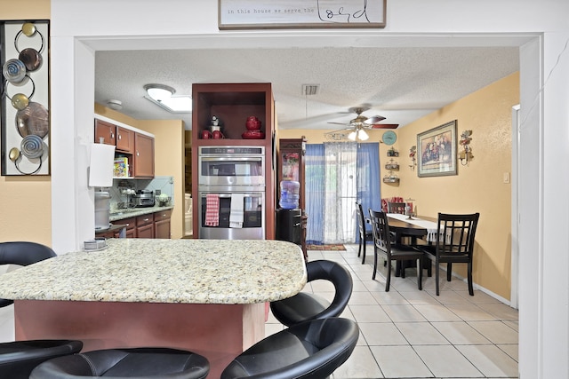 kitchen featuring a textured ceiling, light tile patterned floors, a breakfast bar area, double oven, and ceiling fan