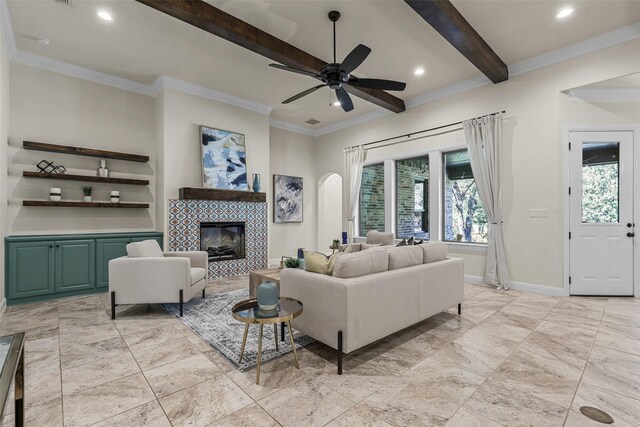 living room featuring ceiling fan, a tiled fireplace, beam ceiling, and ornamental molding