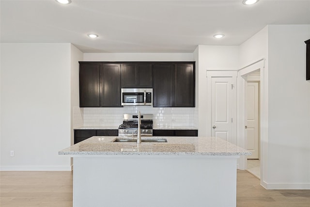 kitchen with stainless steel appliances, a kitchen island with sink, backsplash, and light stone countertops