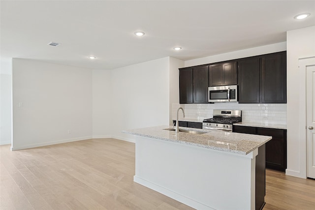 kitchen with light stone counters, light wood finished floors, stainless steel appliances, a kitchen island with sink, and a sink