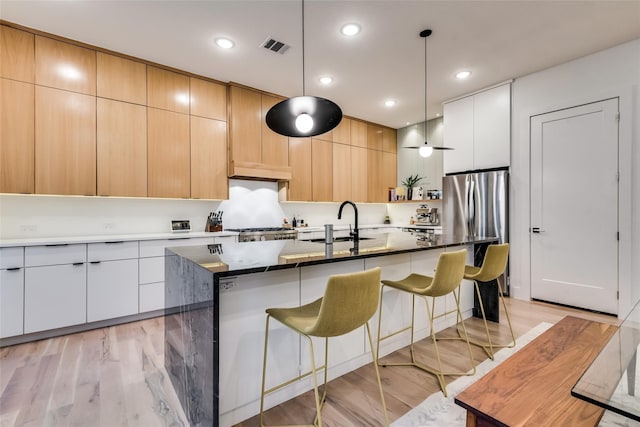 kitchen with stainless steel refrigerator, light hardwood / wood-style flooring, dark stone countertops, an island with sink, and pendant lighting