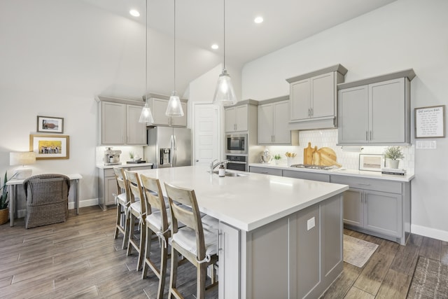 kitchen featuring appliances with stainless steel finishes, a kitchen bar, hanging light fixtures, an island with sink, and gray cabinetry