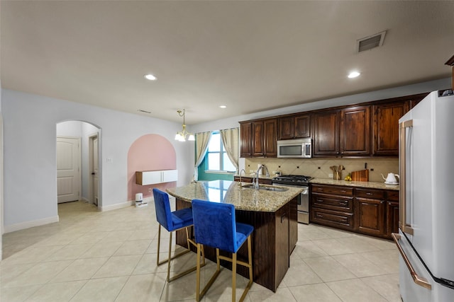 kitchen featuring appliances with stainless steel finishes, light stone counters, a kitchen island with sink, sink, and pendant lighting