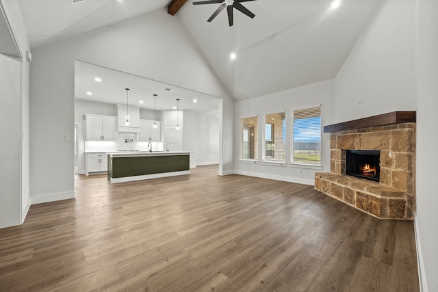 unfurnished living room with ceiling fan, high vaulted ceiling, a stone fireplace, wood finished floors, and beamed ceiling