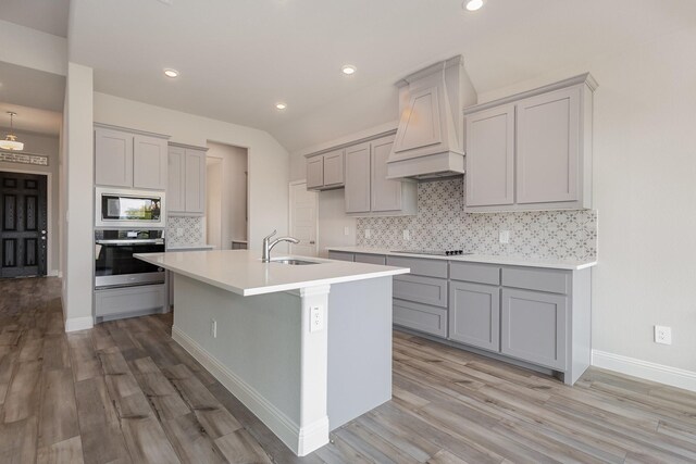 kitchen featuring appliances with stainless steel finishes, backsplash, light wood-type flooring, an island with sink, and premium range hood