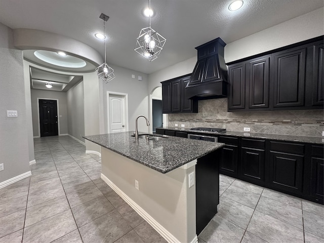 kitchen with tasteful backsplash, a textured ceiling, custom range hood, sink, and a kitchen island with sink