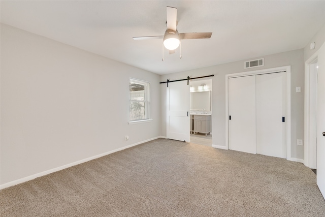 unfurnished bedroom with a closet, a barn door, light colored carpet, ceiling fan, and ensuite bathroom