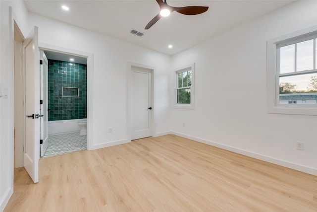 unfurnished bedroom featuring ceiling fan, multiple windows, light wood-type flooring, and connected bathroom