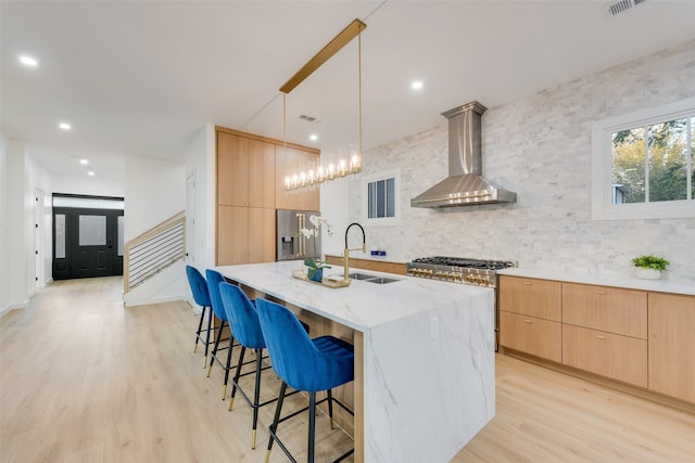 kitchen featuring an island with sink, high quality appliances, wall chimney range hood, light brown cabinetry, and decorative light fixtures