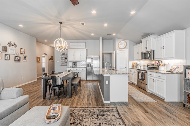 kitchen with a center island with sink, appliances with stainless steel finishes, light stone countertops, hanging light fixtures, and vaulted ceiling