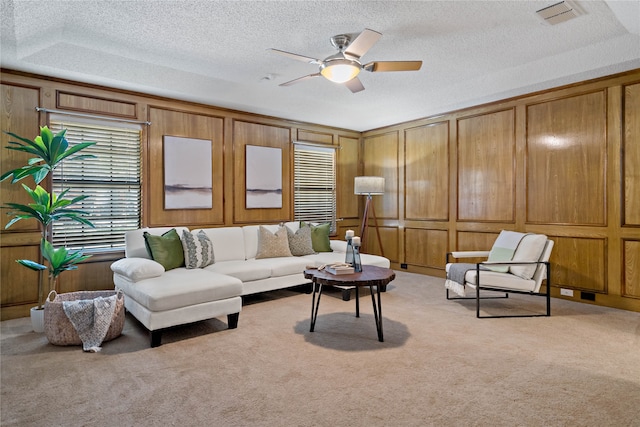 carpeted living room with a raised ceiling, wood walls, ceiling fan, and a textured ceiling