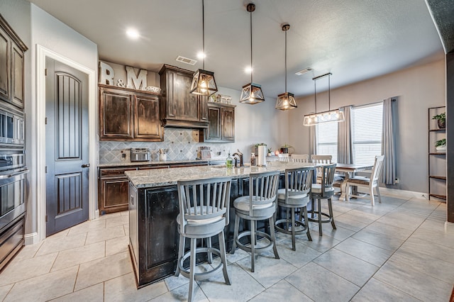 kitchen featuring light stone countertops, a kitchen bar, an island with sink, oven, and pendant lighting