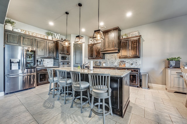 kitchen featuring stainless steel appliances, light stone counters, dark brown cabinetry, an island with sink, and pendant lighting