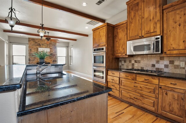 kitchen with stainless steel appliances, beamed ceiling, a center island with sink, decorative light fixtures, and light wood-type flooring