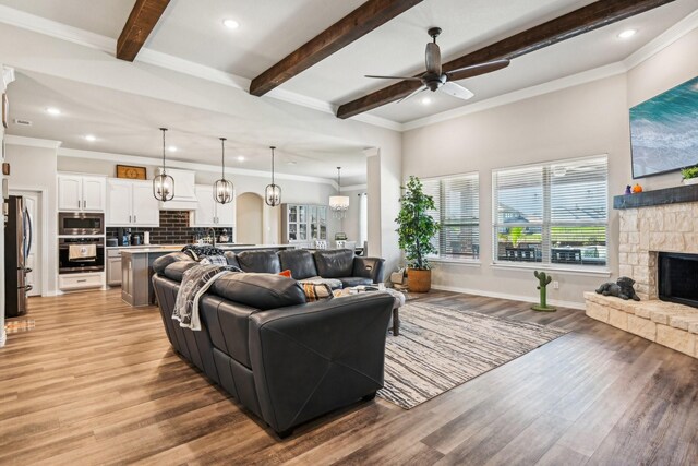 living room with a stone fireplace, light hardwood / wood-style flooring, beamed ceiling, ceiling fan with notable chandelier, and ornamental molding