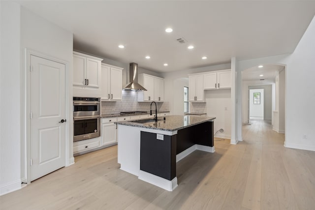 kitchen featuring light hardwood / wood-style floors, dark stone counters, wall chimney exhaust hood, an island with sink, and appliances with stainless steel finishes