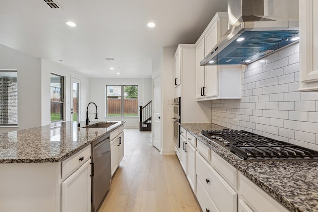 kitchen featuring light hardwood / wood-style flooring, sink, an island with sink, wall chimney range hood, and appliances with stainless steel finishes