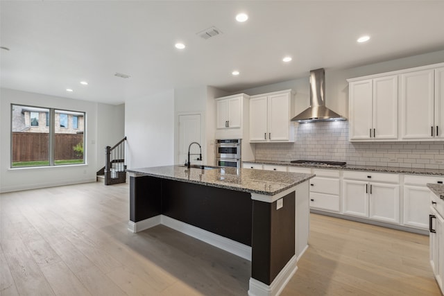 kitchen featuring wall chimney range hood, a center island with sink, light wood-type flooring, and appliances with stainless steel finishes