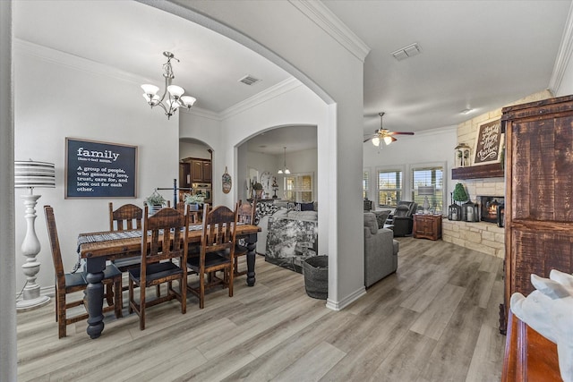 dining area featuring ornamental molding, a stone fireplace, ceiling fan with notable chandelier, and light wood-type flooring