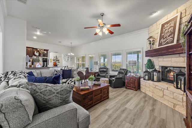 living room with crown molding, a stone fireplace, ceiling fan with notable chandelier, and light wood-type flooring