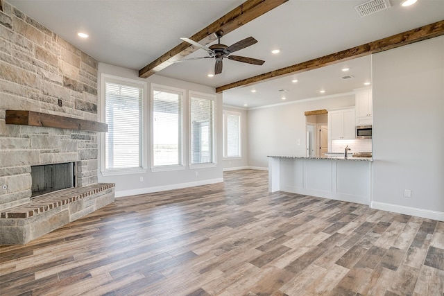 unfurnished living room featuring beam ceiling, a stone fireplace, ceiling fan, and light hardwood / wood-style floors