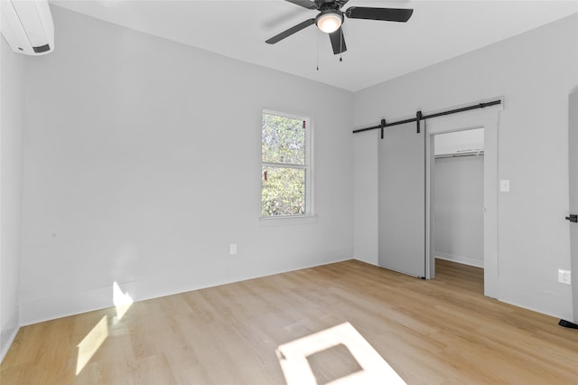 unfurnished bedroom with ceiling fan, a barn door, a wall mounted AC, light wood-type flooring, and a closet