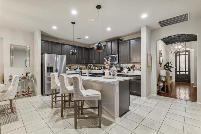 kitchen with backsplash, dark brown cabinets, stainless steel appliances, a center island with sink, and hanging light fixtures