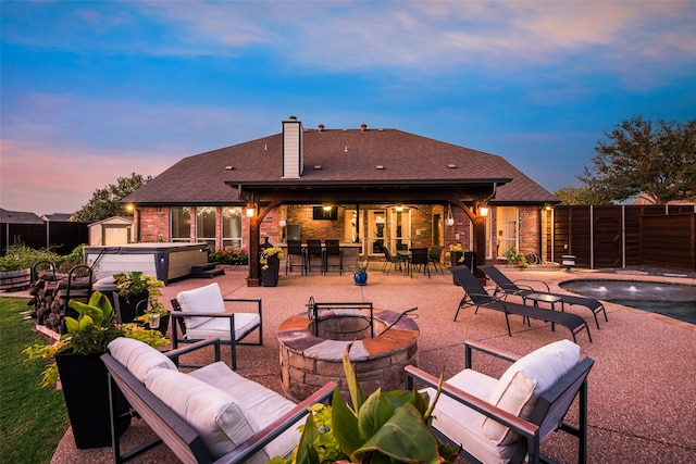 patio terrace at dusk with a pool with hot tub, a bar, and an outdoor living space with a fire pit