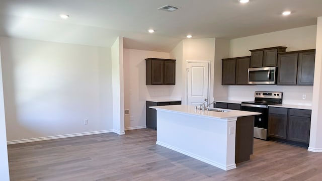 kitchen with sink, appliances with stainless steel finishes, dark brown cabinets, light hardwood / wood-style floors, and an island with sink