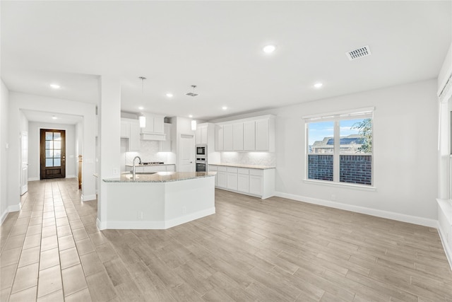 kitchen featuring tasteful backsplash, white cabinetry, hanging light fixtures, a center island with sink, and light wood-type flooring