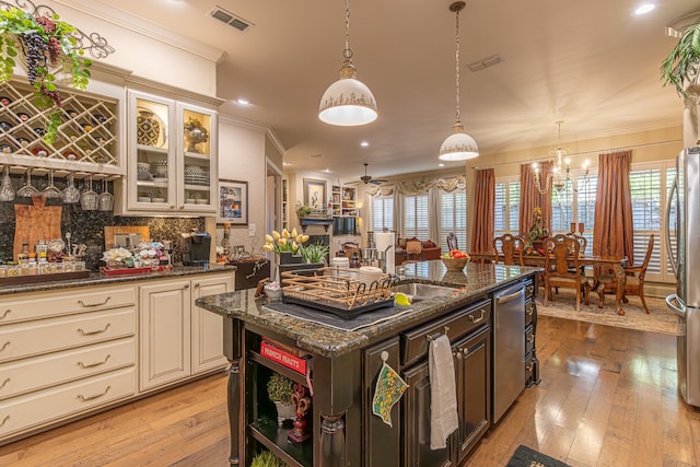 kitchen with ceiling fan with notable chandelier, light hardwood / wood-style floors, dark stone counters, and an island with sink