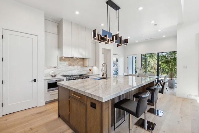 kitchen with pendant lighting, a kitchen island with sink, sink, light wood-type flooring, and white cabinetry