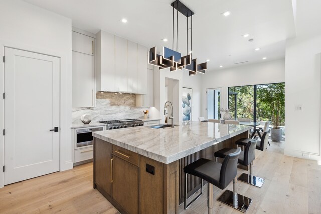 kitchen featuring sink, light stone counters, hanging light fixtures, a center island with sink, and white cabinets