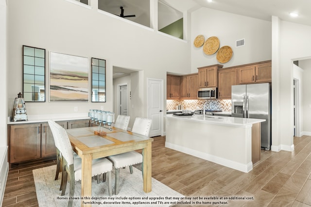 kitchen with appliances with stainless steel finishes, tasteful backsplash, a center island with sink, light hardwood / wood-style flooring, and high vaulted ceiling